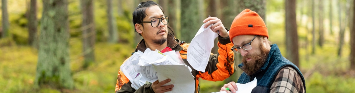 Två män som står med massa anmälningsblanketter mitt i skogen.
